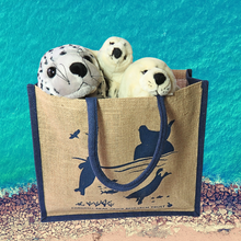 Load image into Gallery viewer, Three seal plushy toys in a jute bag. The bag has navy blue seals on it. The background is of a beach and the sea from above. 