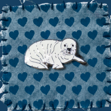 Load image into Gallery viewer, An enamel seal pup pin badge, resting on a blue fabric swatch with a heart pattern.