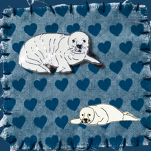 Load image into Gallery viewer, Two enamel seal pup pin badges, one larger than the other. Resting on a blue fabric swatch with a heart pattern.