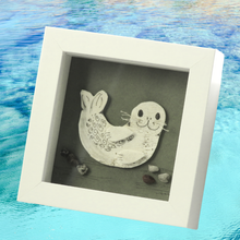 Load image into Gallery viewer, A square white shadow box frame containing a seal artwork on a textured gray background, styled with small stones. The box frame is set against a blue water-textured backdrop.