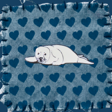 Load image into Gallery viewer, An enamel seal pup pin badge, resting on a blue fabric swatch with a heart pattern.